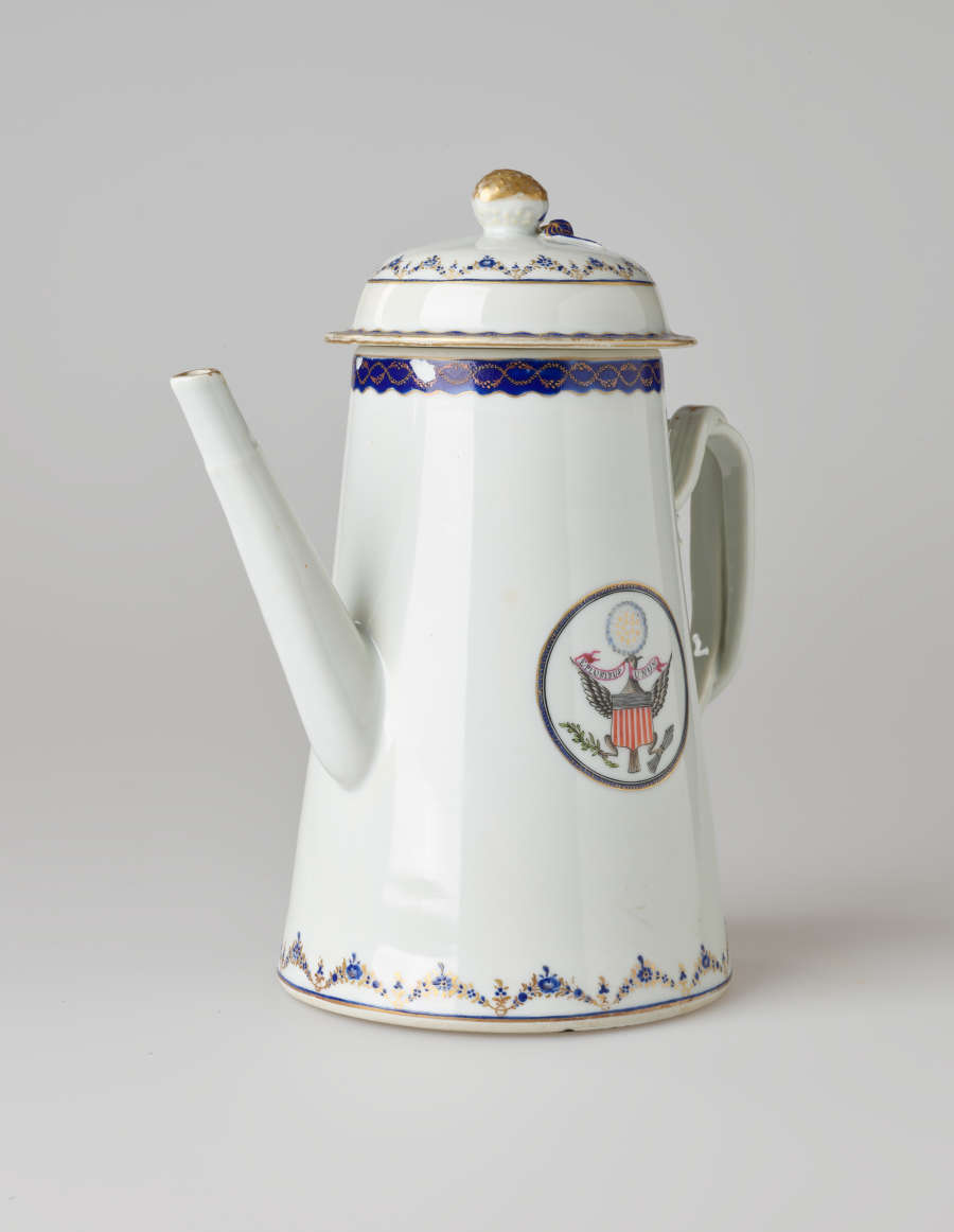 A white coffee pot with dark blue and gilded decorations, a straight spout, angular handle, and heraldry imagery in a circle on the body of the coffee pot.