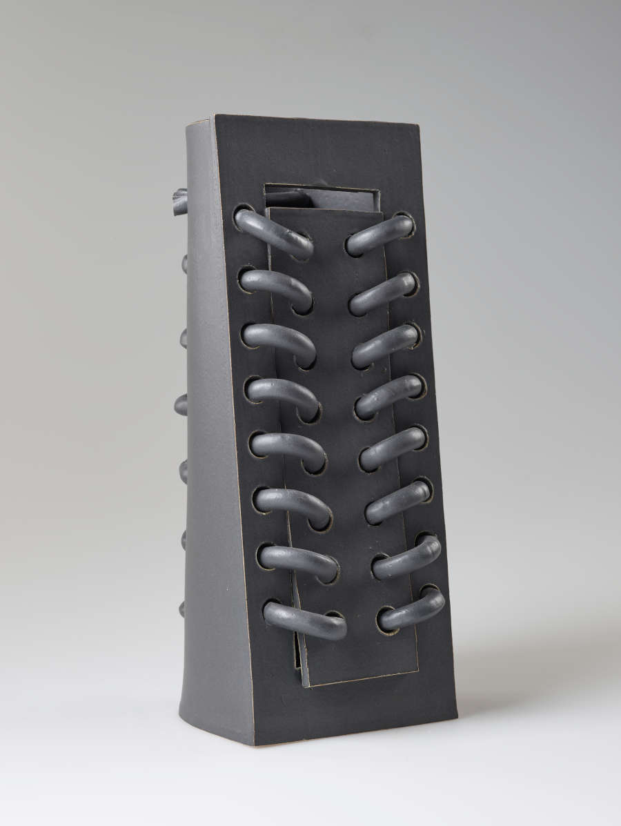 Diagonal front view of a rectangular gray ceramic sculpture featuring a row of horizontal loops threaded through and protruding from the surface.