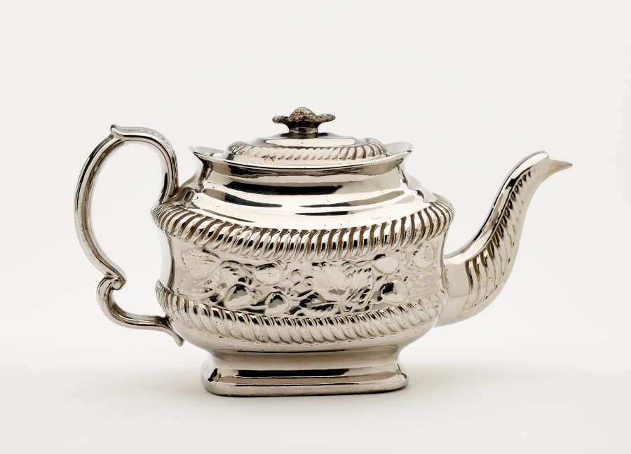 A silver teapot with a decorative handle, a rounded square body and spout with sculptural decorations.