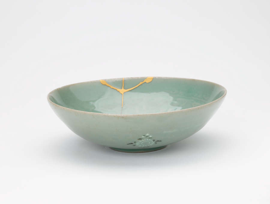 Side-view of a tilted, light-teal, stout glazed bowl, with green-blue floral motifs, faded floral patterns, and gold patched cracks along a top-edge.