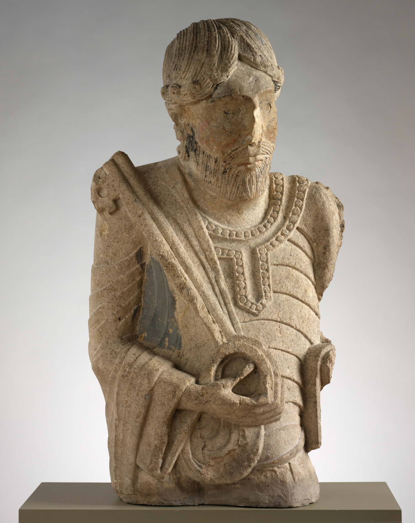 Stone sculpture of a bearded man with curled hair, one hand grasping a key, the edges softly weathered.