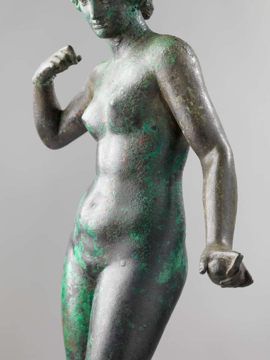 A tarnished bronze statue of a standing nude woman gripping an object in one hand and curling her arm towards her shoulder, looking slightly downwards.