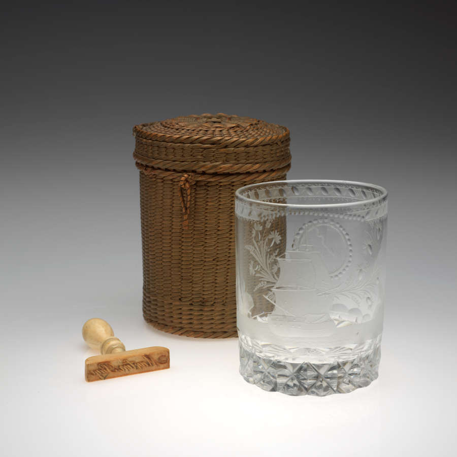 Rounded clear glass with etched designs next to a brown woven basket of the same shape. A small, tan-colored implement lays to the left of the basket and glass.