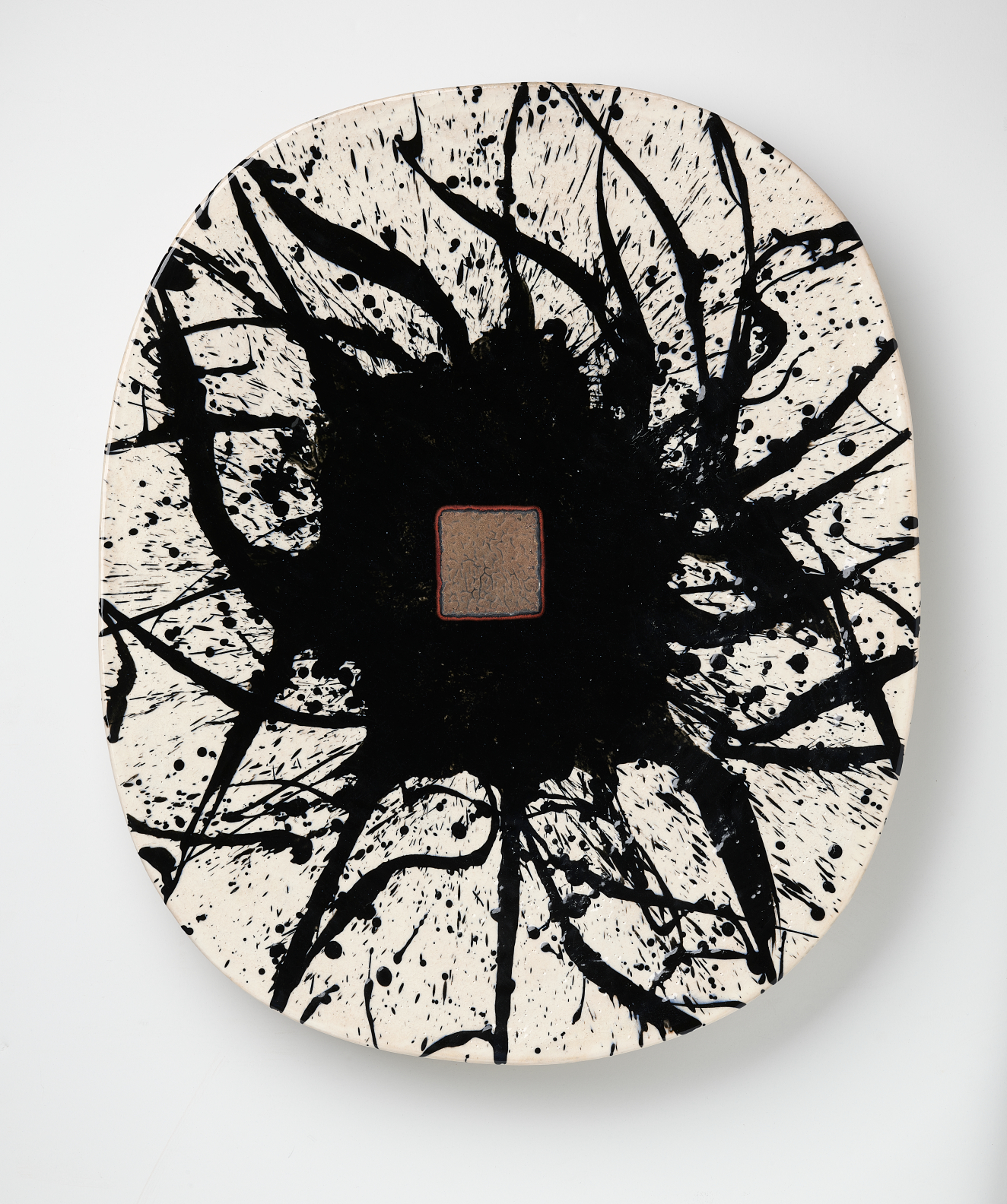 A flat oval ceramic platter featuring an explosive black ink splatter radiating outward from a small central brown rectangle, contrasting with the pure white ground.