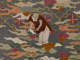 Embroidered detail depicting a bald figure wearing a brown robe and white pants holding a ritual object and bending down atop a backdrop of flowers and swirling clouds.