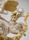 Back view of a silver cherub with curly hair and small, gold wings, one arm holding the end of gold garland while perched up a star-studded robed figure.