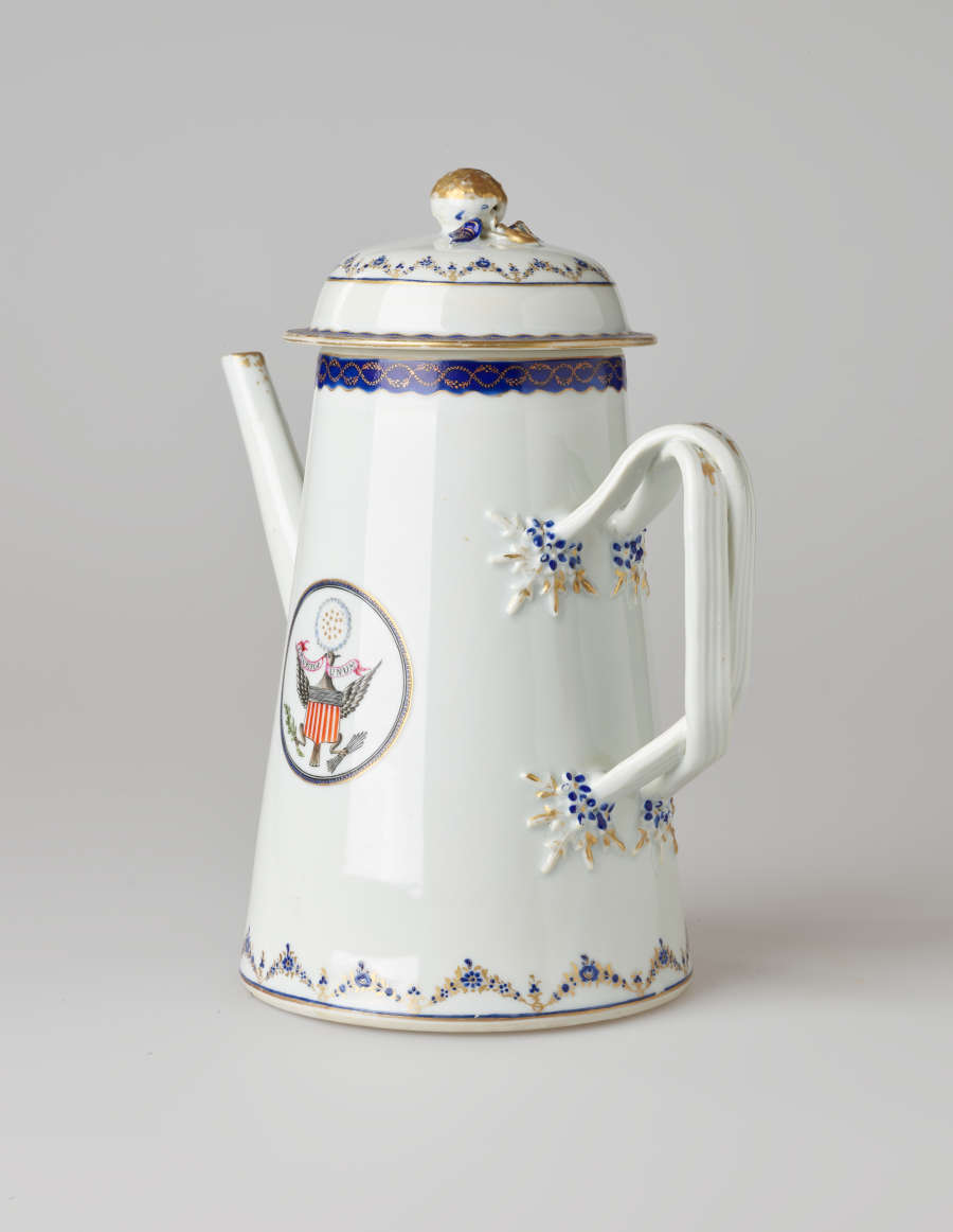 A white coffee pot with dark blue and gilded decorations, a straight spout, angular handle, and heraldry imagery in a circle on the body of the coffee pot.