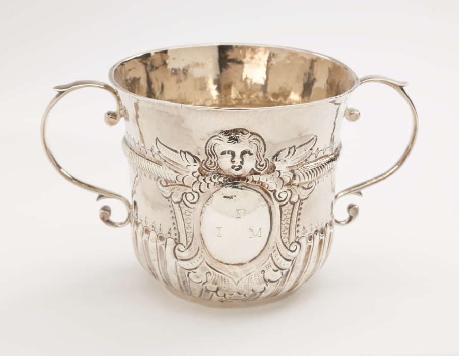 A two-handled silver cup. Incised image of a stylized angel head with wings above an oval shape containing the monogram “P I M.”