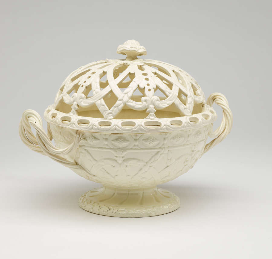 A cream-colored basket with sculptural woven handles, sculpted foot, and a lid with cutout designs.