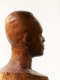 Close-up, three quarter back view of a textured, but glossy, wooden-carved bust depicting a male figure. The sculpture is dramatically lit, casting the face in shadow, while still retaining detail.