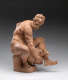 Realistically carved terracotta sculpture depicting a muscular bearded man sitting and leaning over to hold a vessel between his legs, one of which is wrapped over top of it.