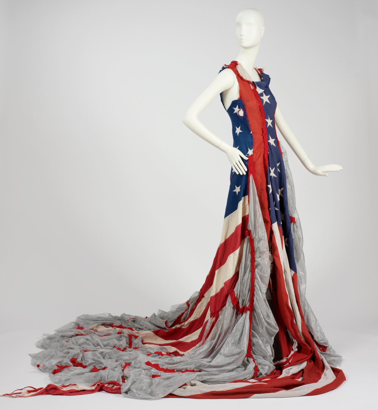 Sleeveless dress designed from a red, white, and blue stars-and-stripe patterned material resembling the American flag with sheer grey fabric cascading to the floor in a sweeping train. 