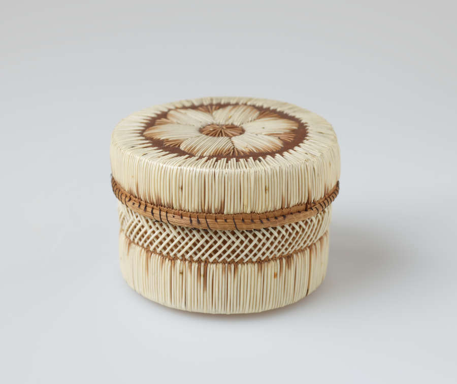 Cylindrical lidded leather box with cross-hatching and linear stripe patterns woven in white and brown straw. The brown rimmed lid has a central floral motif woven atop it.