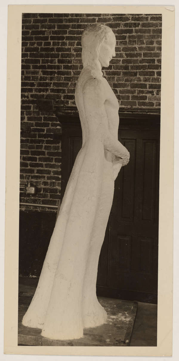 Photograph depicting a sculpture of a figure leaning forwards; a brick wall is visible in the background. The figure is in drapery and the hands are placed above the pelvis.