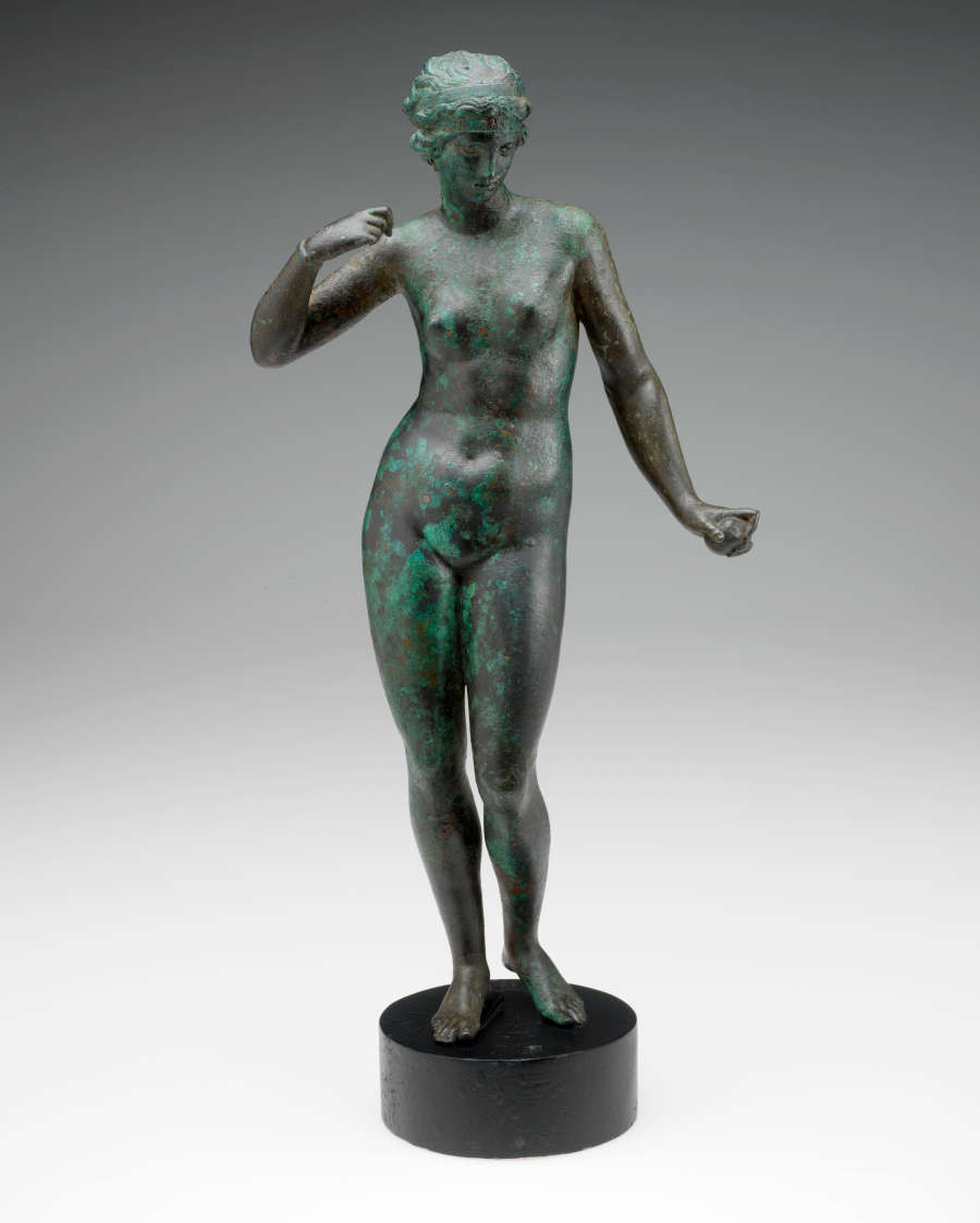Gray metal sculpture with a green patina of a crowned nude woman standing on a circular pedestal, one arm down, grasping an object, and the other curled towards her shoulder.