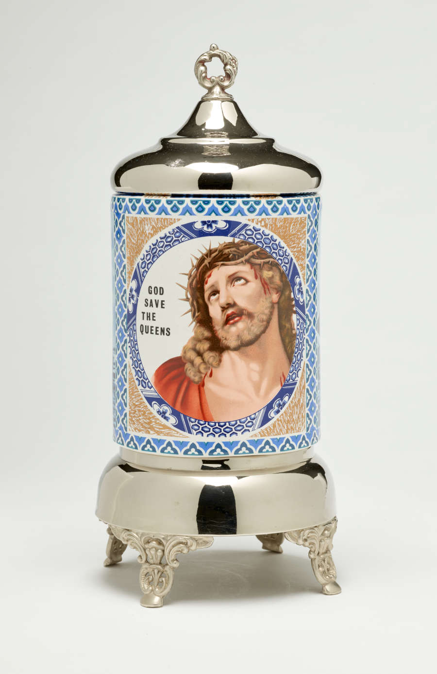 Sculpture with ceramic body covered in blue, white, and tan decoration. Image in the center of Jesus next to the words “God save the queens.” Ceramic is framed by a metal top and base on four ornate feet.