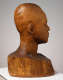 Three quarter back view of a wooden bust depicting the head of a male figure. His expression is stern and holds an intense gaze. The wood is cracked, retaining natural grain.