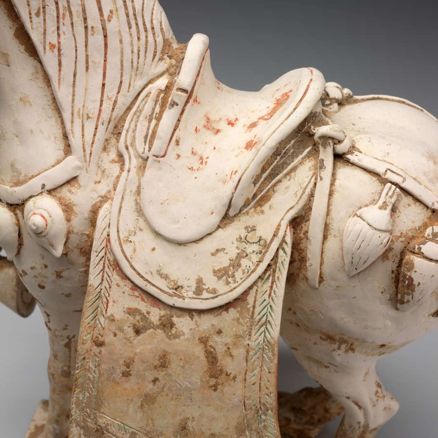 Close-up of the intricately sculpted layered saddle of a ceramic sculpture of a horse along with its etched mane and ornamental tassel belts. Remnants of its painted surface are visible.
