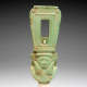 Green glazed amulet with slightly worn brown edges of a carved face, calm expression, smooth long hair, and wide rounded neckline wearing a tall headpiece with rectangular hole. 