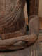 The hands of a wooden Buddha statue resting in its lap, with its fingers forming a circle with the weathered wood showing deep cracks and rough textures.