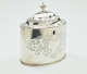 A silver tea caddy with a lid and sculptural finial. There are swirling engravings along the body and lid.