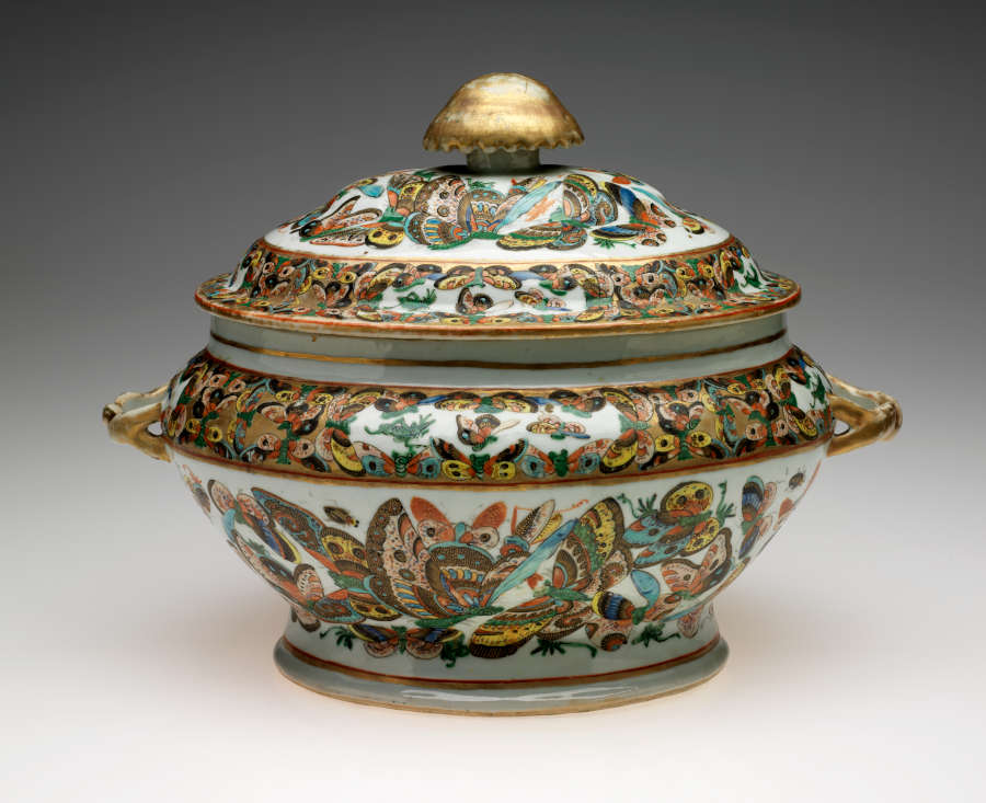 An oval lidded vessel with a large gold finial. The body of the vessel is decorated with multicolored butterflies on a white ground.
