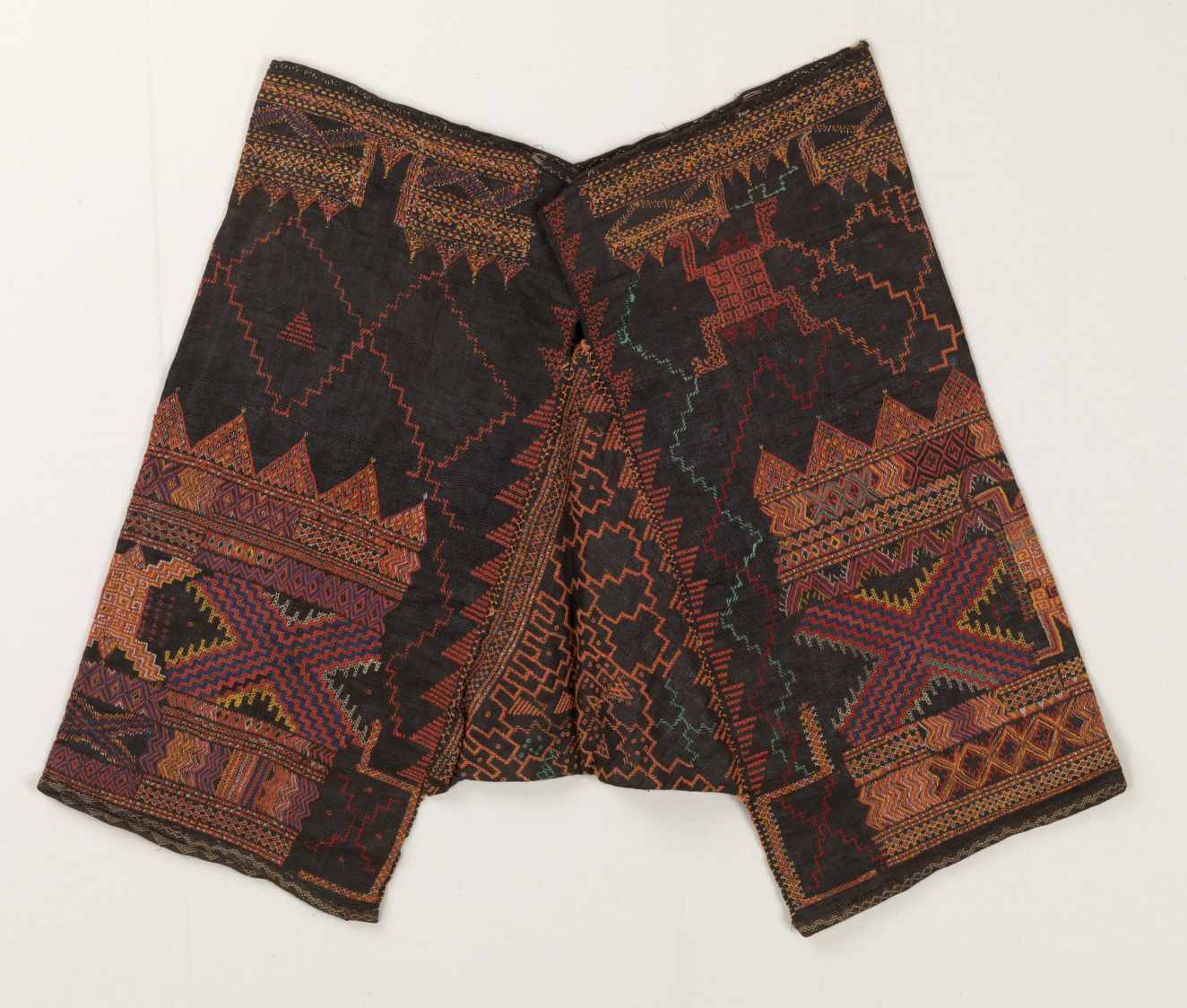 Pants made of dark fabric on a light background adorned by intricate embroidery in earth tones. Each leg has a large X-shape in purple and orange surrounded by triangular motifs.