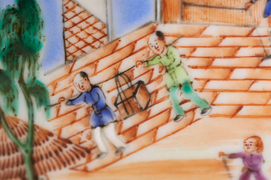 A close up of a painted ceramic depicting two figures dressed in blue and green using a shoulder pole to carry a box, with a tree and brick pathway.