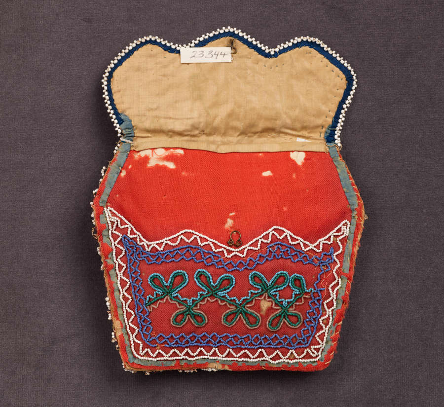 Red irregular hexagonal bag with an open jagged flap, tan on the inside. Wavy triangular patterning in whites and blues are beaded around the border with green knot-like motifs inside.