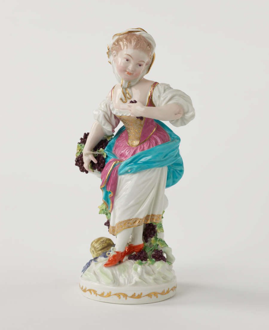 Sculpture of a light skinned figure with grapes dressed in historical clothing that is gold, white, blue, pink, and red.