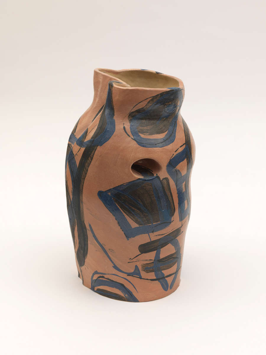 Asymmetrical clay vessel in warm pink tones with a small opening on its side, its uneven rim and subtly lopsided form painted with abstract blue and black geometric designs.