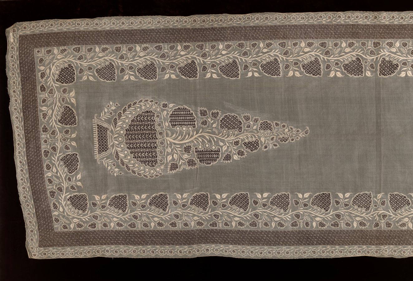 Rectangular transparent fabric on a dark background. Features floral embroidery patterns which form a cone-shape surrounded by an intricate border made up of darker and lighter sections.