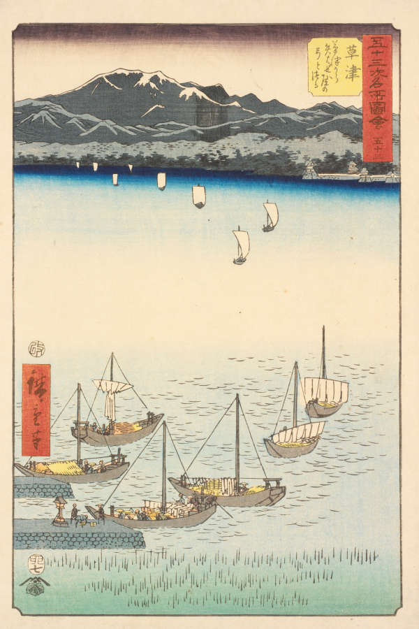 Several sailboats carrying cargo float on calm blue waters, with distant snowy mountains and a tranquil horizon.