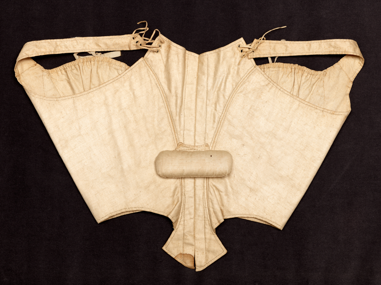 An aged cream-colored corset laid flat featuring structured boning, side shoulder straps, laced ties, and a padded cylinder inserted at the center. 