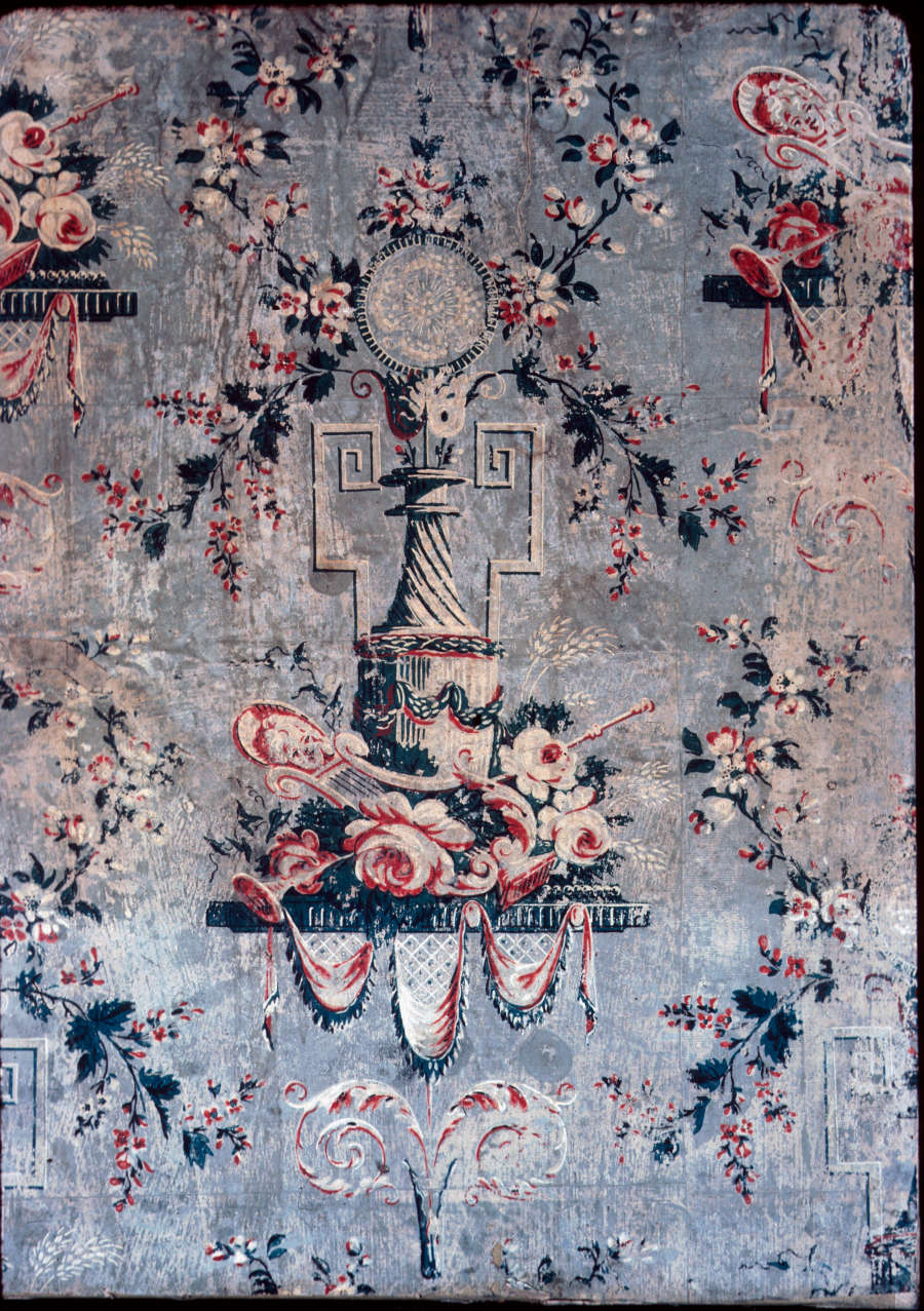 Weathered print of red and white flower garlands surrounding a lined center pillar which holds a circular pendant above. Instruments, flowers, and draping fabric are at the object’s base.