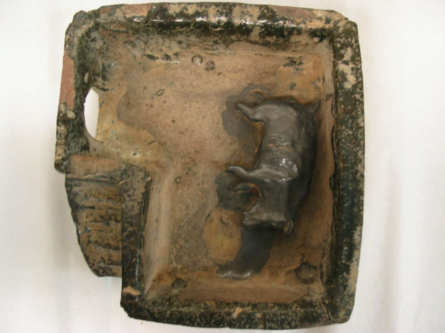 Ceramic sculpture showing a small pig laying on its side, enclosed by a rectangular structure which features arches and stairs. The sculpture is tan with a glossy muted green glaze. 