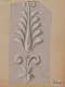 A light gray ornament, featuring a symmetrical leaf pattern above a floral design with curled accents.