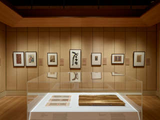 Gallery space with tan walls and peaceful lighting. Several works hang on the far wall, with additional objects in a case in the foreground.
