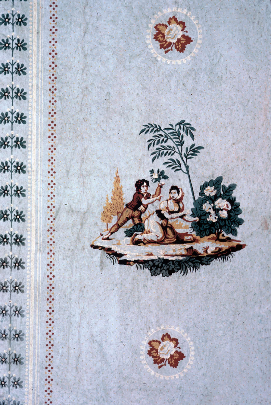 Closeup of a tapestry’s edge showing two figures picking flowers. A green floral border lines the outermost edge, followed by a red dotted border which repeats on both sides.