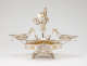 Elaborate silver double-oval-tray centerpiece with central winged-figurine holding up a gold garland with two cherubs, framed by swirling arms extending to two shallow dishes, all elevated on four ornate legs.