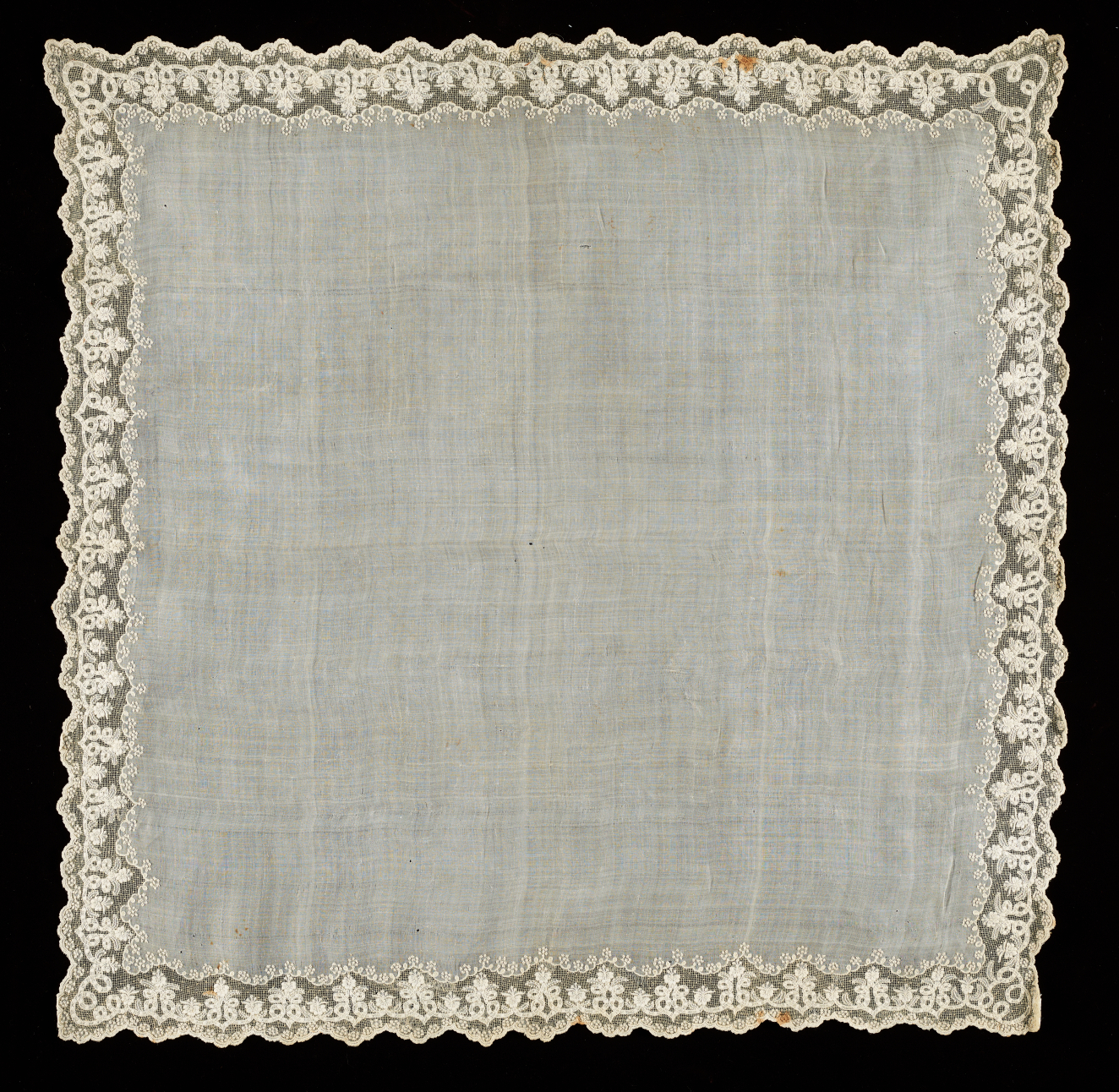 Square light colored fabric with slight transparency on a black background with an intricate lace border. The border features delicate embroidered patterns in contrast to the undecorated center.