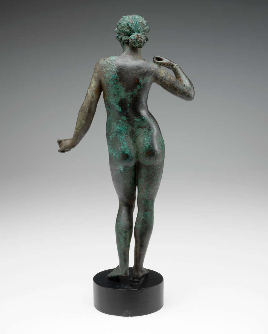 Back view of a tarnished bronze statue of a standing crowned nude woman with one arm curled towards her shoulder and the other held out, standing on a circular pedestal.