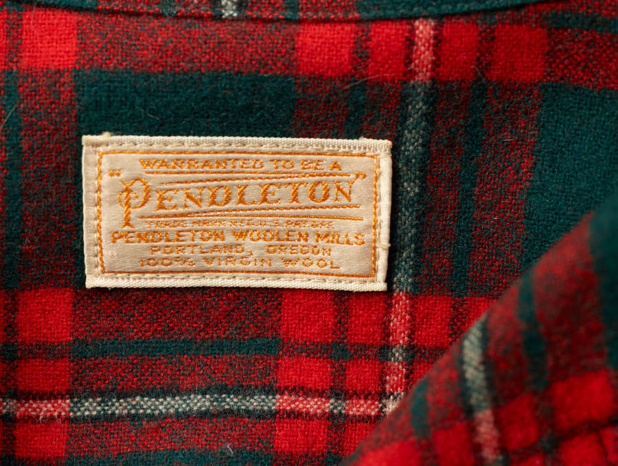 Close-up of a tan rectangular label with orange embroidered text that reads “warranted to be a ‘Pendleton.’ Pendleton Woolen Mills. Portland, Oregon 100% Virgin Wool, ” on a red and green plaid long-sleeved shirt. 
