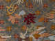 Intricate embroidery on a gray fabric, depicting a golden Chinese character above a deep red character an old man on his back points up at, atop a backdrop of flowers. 