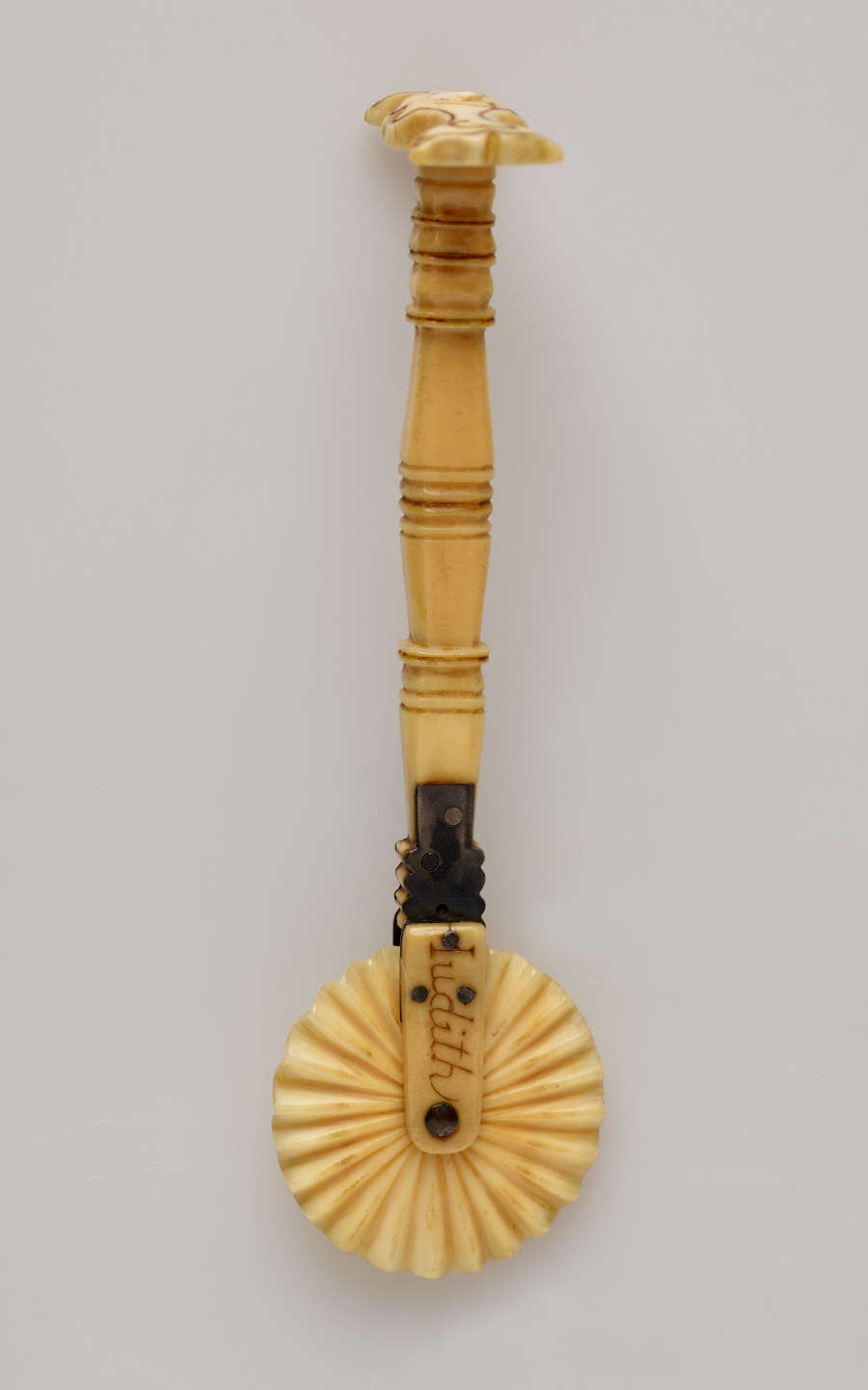 A tan colored tool with decorative handle and a textured wheel. There are letters on the part attaching the wheel to the handle.