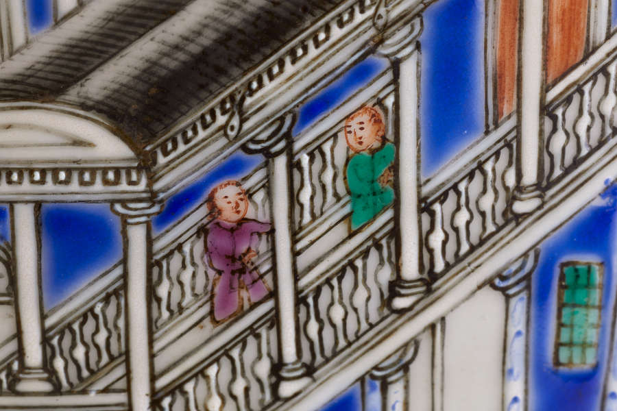 A close-up of a painted ceramic depicting two figures dressed in purple and green walking along a decorative bridge with detailed railings.