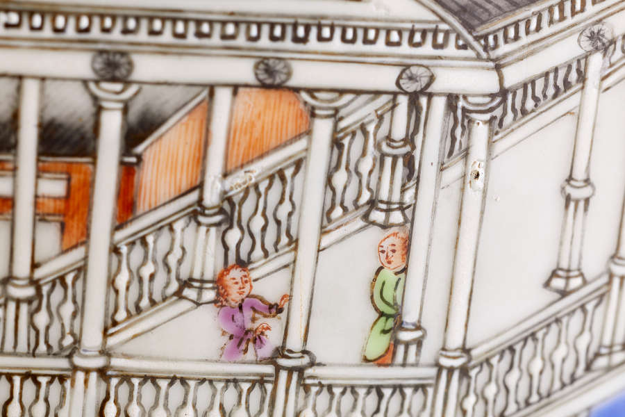 A close up of a painted ceramic depicting two figures dressed in purple and green, standing on an ornate balcony with detailed railings and columns.