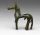 Side-view of a bronze horse figurine with a green patina, and an exaggeratedly thin torso, thick neck, long legs and tail and long narrow snout.