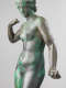 Cropped image of a gray metal sculpture with a green patina of a standing crowned nude woman, one arm down, grasping an object, and the other curled towards her shoulder.