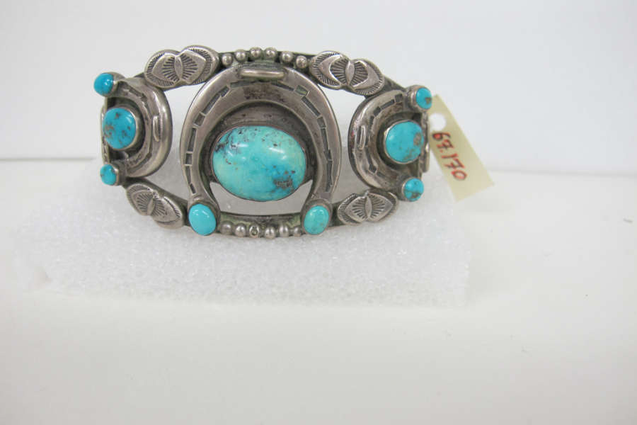 Silver and turquoise bracelet on a rectangular white base. The bracelet features two rows or ornately patterned silver connected by turquoise stones enveloped by silver crescents. A yellow label reads “67.170”.